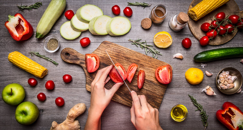 Uma bancada de cozinha com vários alimentos espalhados e uma tábua ao meio na qual alguém corta tomates